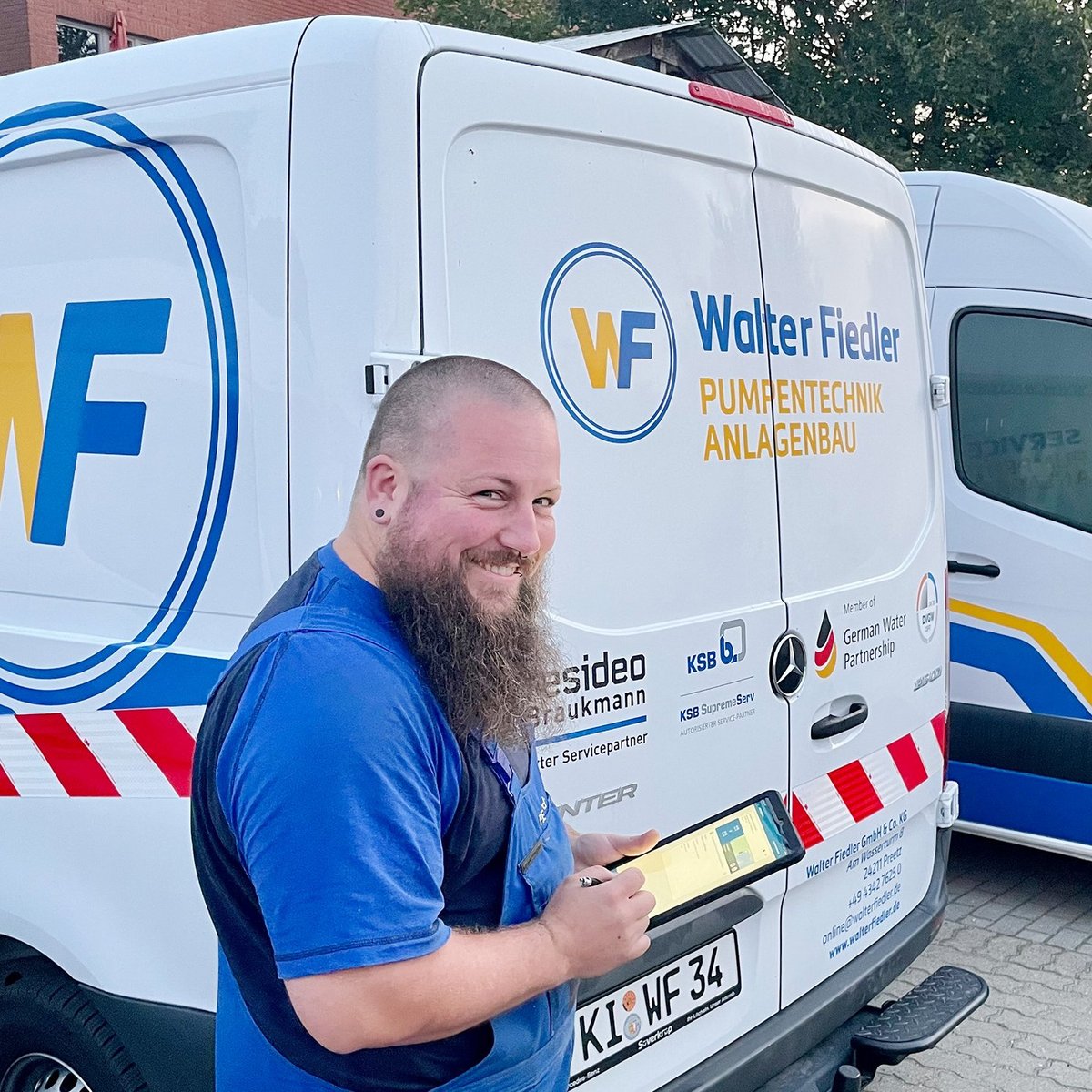 Der Mann in blauer Arbeitskleidung lächelt, während er an einem Lieferwagen mit dem Logo 'Walter Fiedler' steht.