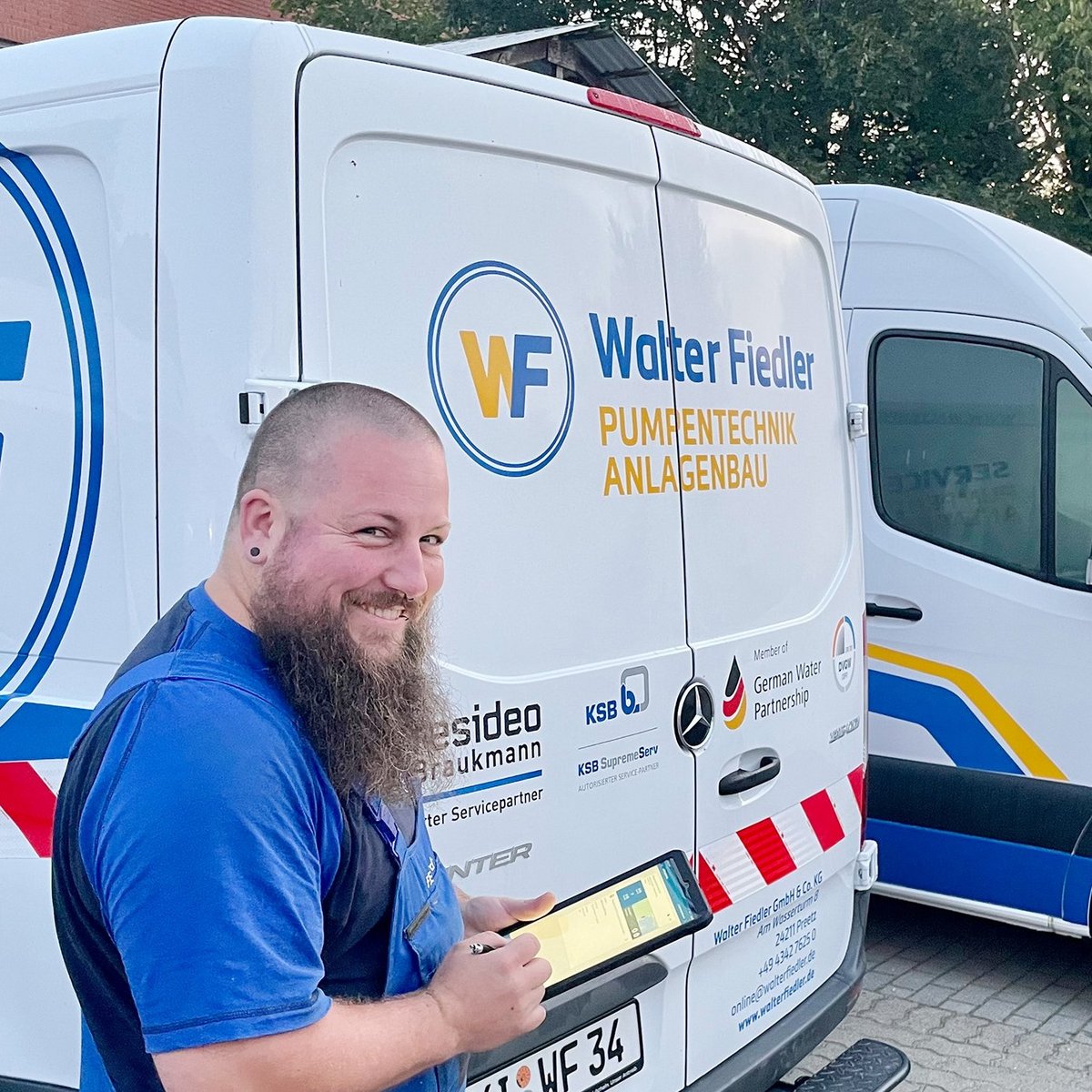 Der Mann in blauer Arbeitskleidung lächelt, während er an einem Lieferwagen mit dem Logo 'Walter Fiedler' steht.