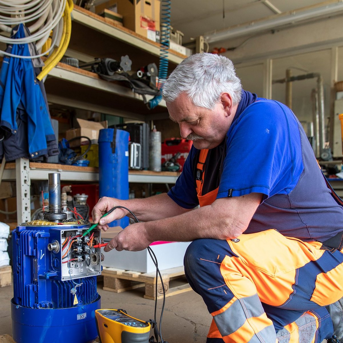 Ein Mann in orangefarbener Arbeitskleidung repariert einen Elektromotor in einer Werkstatt, umgeben von Werkzeugen und Materialien.