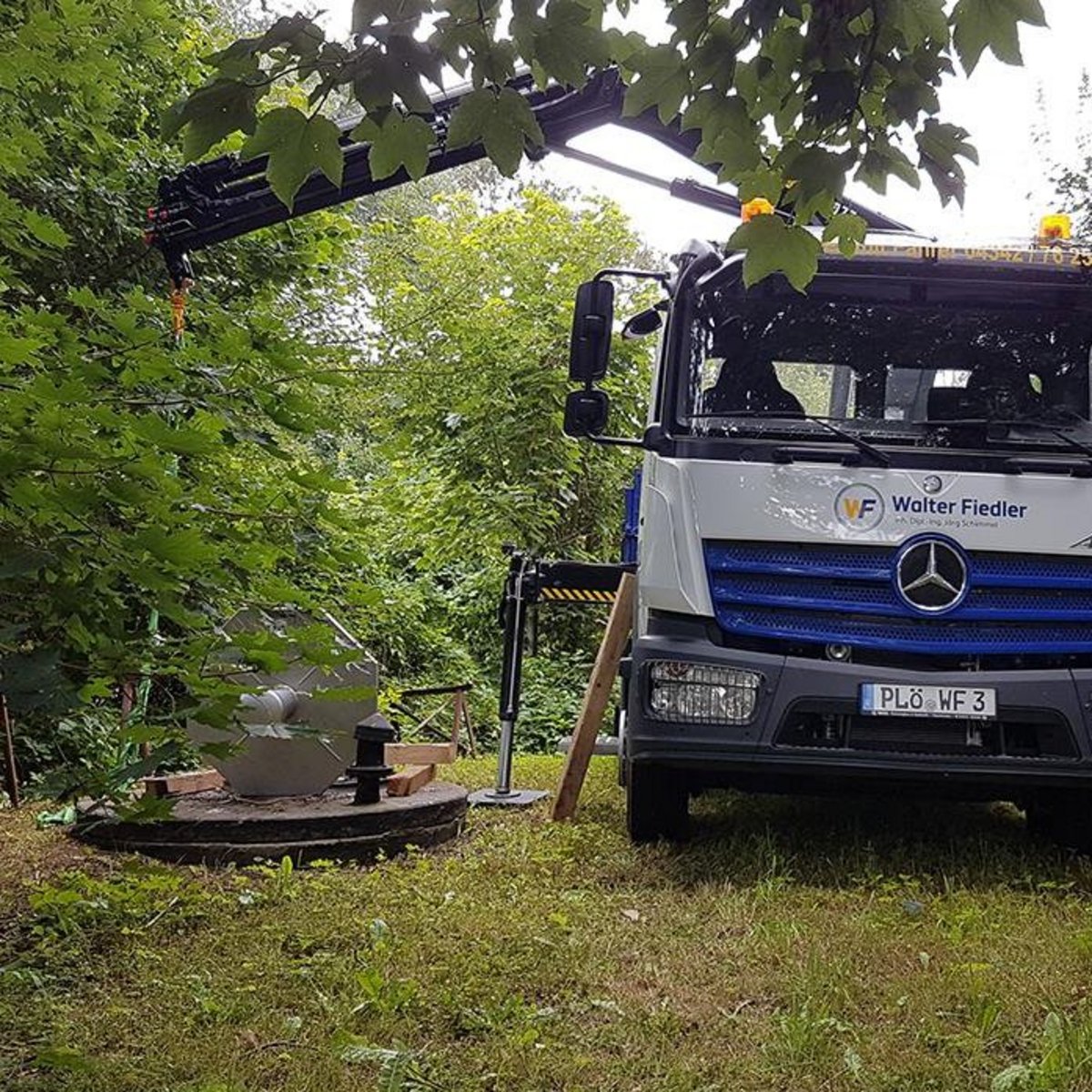 Der Lkw von Walter Fiedler ist in einer grünen Umgebung geparkt, während ein Kranarm bereit ist, eine Last zu heben.