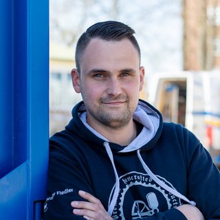 Ein junger Mann mit einem lässigen Hoodie steht entspannt an einer blauen Wand und lächelt selbstbewusst in die Kamera.