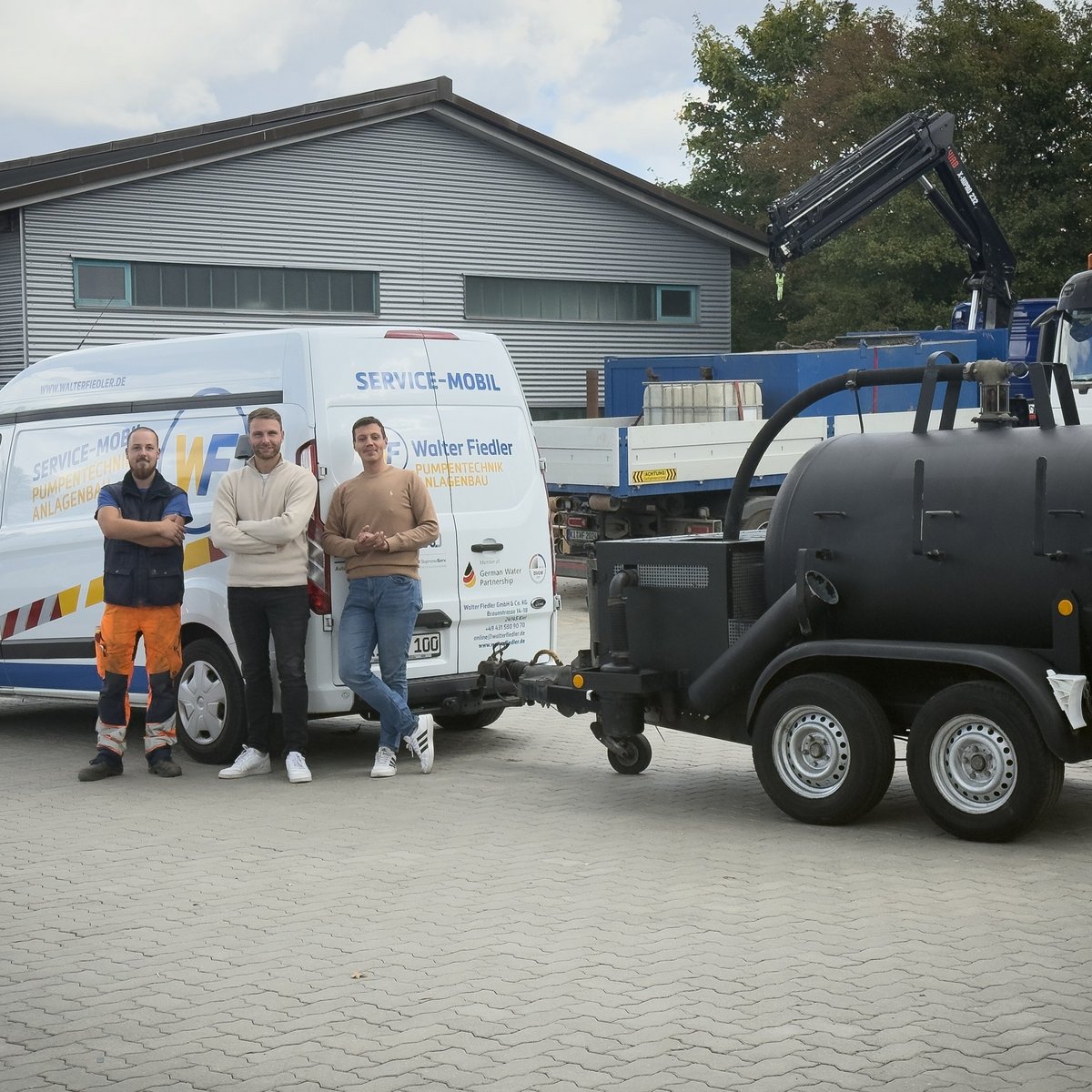 Die Szene zeigt drei Fachleute in Arbeitskleidung, die stolz vor ihrem Service-Mobil und einem speziellen Anhänger stehen.