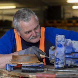 Ein älterer Mann mit grauen Haaren und einem Schnurrbart arbeitet konzentriert an einer blauen Maschine in einer Werkstatt.