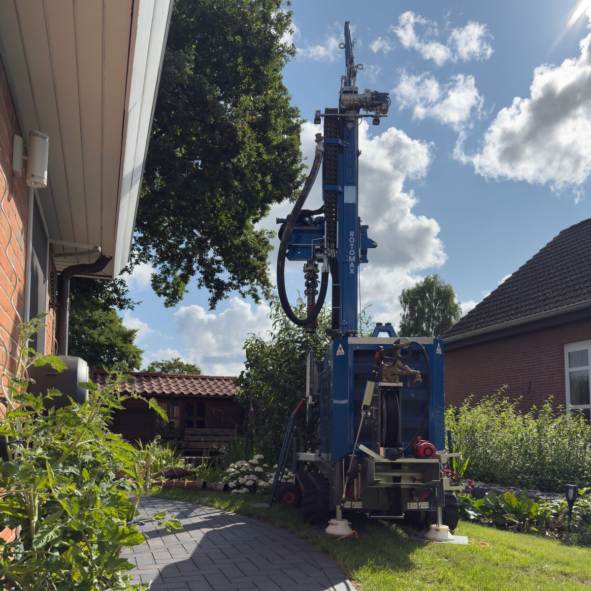 Ein gepflasterter Gartenweg führt zu einem blauen Bohrgerät, das unter einem strahlend blauen Himmel arbeitet.